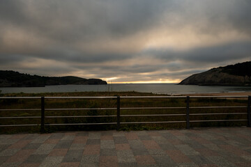 Sc view of a waterfront with hills at sunset, featuring a railing and a paved walkway in the foreground..