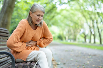 Elderly woman sitting on park bench outside experiencing stomach discomfort. She appears to be in...