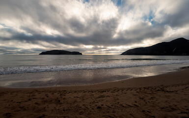 Cal beach with gentle waves, distant islands, and a dramatic, cloudy sky at sunset....