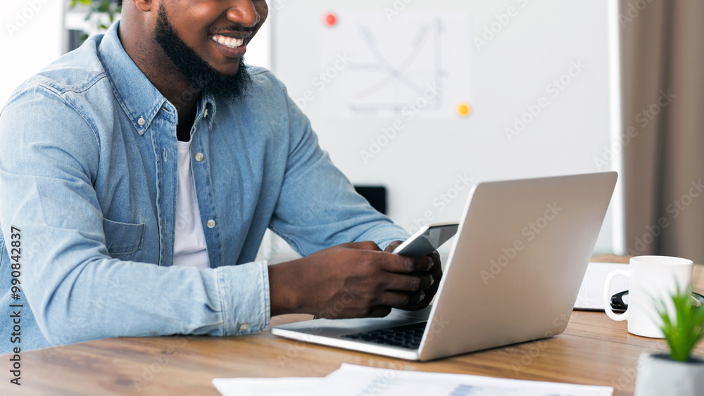 Poster Modern technologies at work. Smiling afro entrepreneur using smartphone and working on laptop in office.