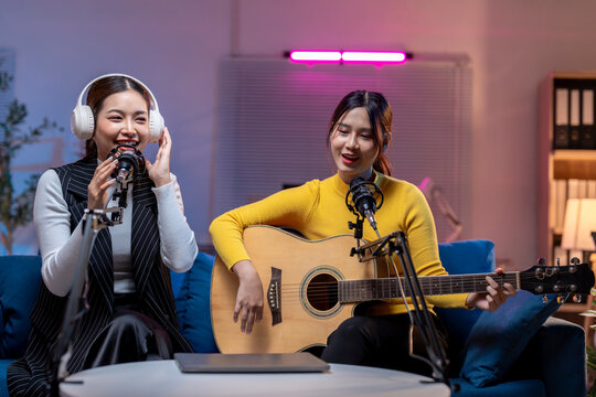 Two asian women playing guitar and singing into microphone recording podcast