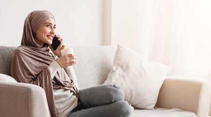 Home Rest. Young Muslim Girl In Hijab Drinking Coffee And Talking On Cellphone, Relaxing On Sofa In Living Room, Free Space