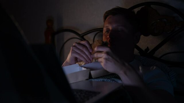 Hungry young man relaxing evening at home, lying comfortably in bed while watching movie on laptop and eating tasty fast food. Happy tired male enjoying eating burger in bed while using computer.