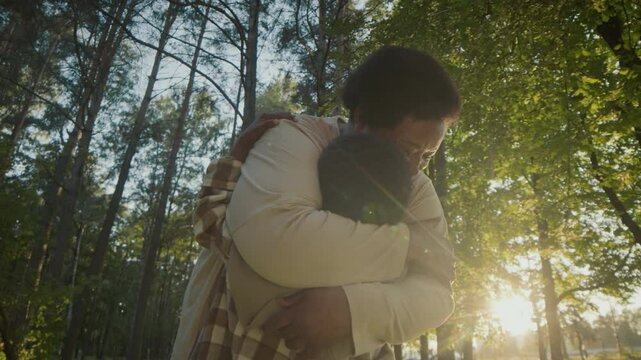 Low angle full shot of African American senior woman lovingly embracing and kissing grandson on cheek during walk in forest on warm sunny day. Black boy hugging and giving high-five to grandmother