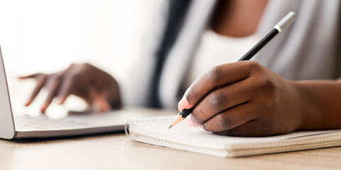 Unrecognizable black female employee taking notes while working on laptop in office, selective focus on arm with pencil