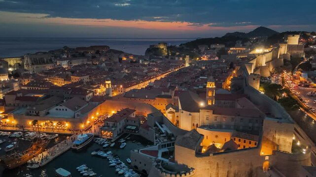 Dubrovnik walls and old town after sunset with city lights. Aerial shot of historic city of Dubrovnik in Croatia, UNESCO World Heritage site. Illuminated streets, buildings and marina with boats
