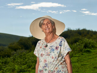 An octogenarian woman with a sun hat poses in front of a meadow with a mountain in the background...