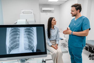 Caucasian young male medic doctor specialist radiologist in blue uniform explaining diagnosis on lung x-ray fluorography to female patient during medical appointment in hospital.