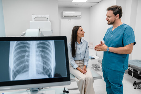 Caucasian young male medic doctor specialist radiologist in blue uniform explaining diagnosis on lung x-ray fluorography to female patient during medical appointment in hospital.