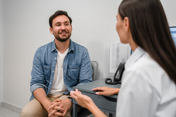 Fototapeta premium Smiling woman doctor listening to male patient complaints at meeting in hospital. Friendly female therapist physician wearing white uniform consulting young man at medical appointment