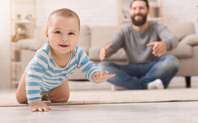 Play with me. Adorable baby boy crawling on floor at home, daddy watching his son, copy space