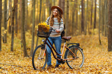 Fototapeta premium Young woman smiling while pushing a bicycle filled with colorful autumn leaves on a sunny day in the park. Active tourist enjoying sunny weather. Weekend concept.