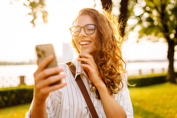 A beautiful woman with curly hair smiles while using her smartphone in a sunlit park by the water...