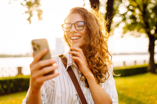 A beautiful woman with curly hair smiles while using her smartphone in a sunlit park by the water during the golden hour. Technology, connection, blogging,