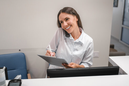 Female receptionist talking to patient on mobile phone, smiling, using computer at workplace, telling healthcare checkup test results, bringing good news, using modern technology for communication