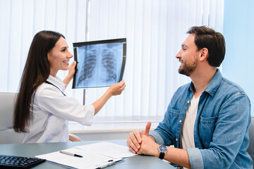 Lung disease. Young female doctor showing chest x-ray to her male patient man at table in modern clinic explaining diagnosis, discussing scan results in hospital office while appointment.