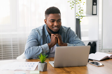 Naklejka premium Thinking about future success. Pensive black businessman looking at laptop screen at workplace in office.