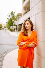 A woman wearing a vibrant orange dress strolls through an elegant city plaza, thoughtfully tucking her hair behind her ear as she admires the surroundings. Concept of lifestyle, fashion, travel.