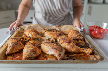  Woman with apron serving fresh and homemade cooked roast chicken legs on a baking tray in the kitchen