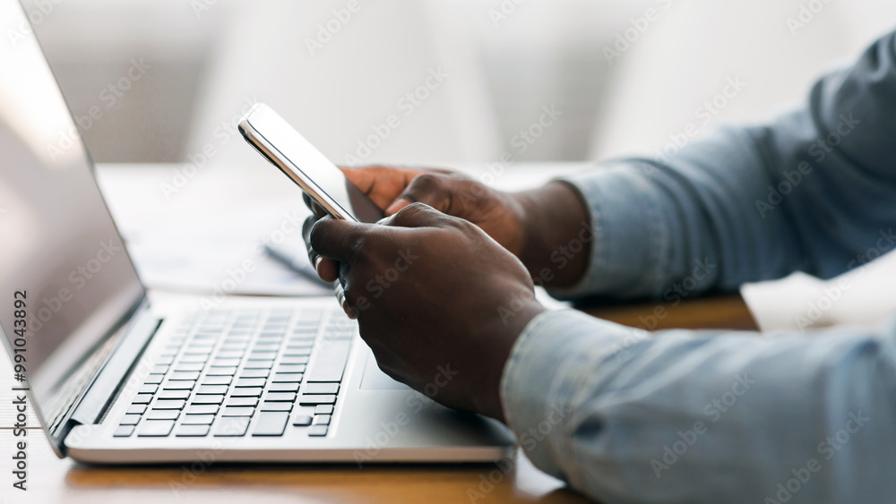 Poster Unrecognizable afro businessman messaging on smartphone and working with laptop in office, side view with copy space