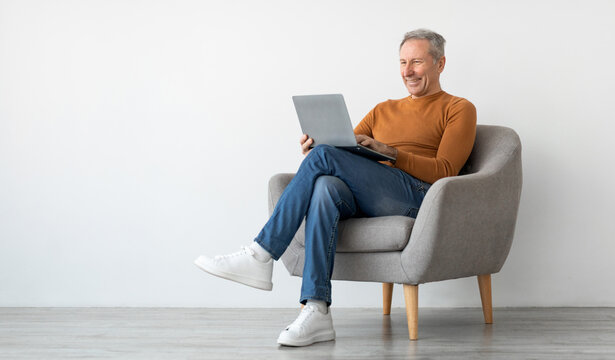 Smiling mature man using laptop sitting on armchair in living room. Happy confident senior male adult resting at home working on pc, typing on keyboard isolated on white studio wall, full body length
