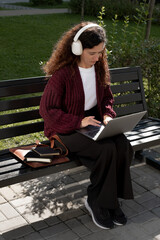 Adult student working remotely on laptop outdoors in headphones. Freelancer woman with laptop and headphones working outside on park bench