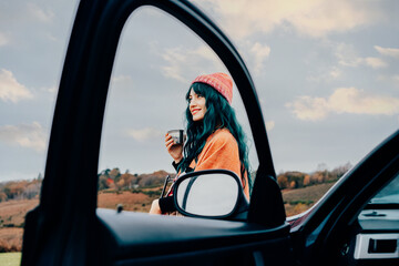 Hipster woman enjoying warm drink on car hood while taking in serene rural landscape during cold season road trip. Female wrapped in Shawl Poncho Cape and hat. Fall, winter travel, vacation concept