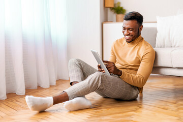 Weekend Concept. Cheerful Afro Guy Using Digital Tablet And Smiling Sitting On Floor At Home. Selective Focus