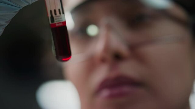 Close-up view with focus on foreground of female lab scientist in goggles and gloves examining blood in test tube