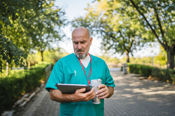 Fototapeta premium Mature senior man doctor use tablet and drink coffee in front hospital