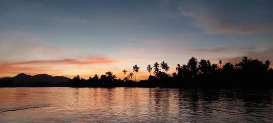 Serene sunset over a peaceful river with silhouettes of trees and distant mountains captured in a tranquil evening scene.