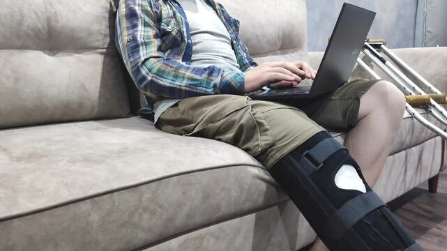 man with orthosis on broken leg sits on sofa and works on laptop. Work from home during illness and recovery, recovery after leg surgery