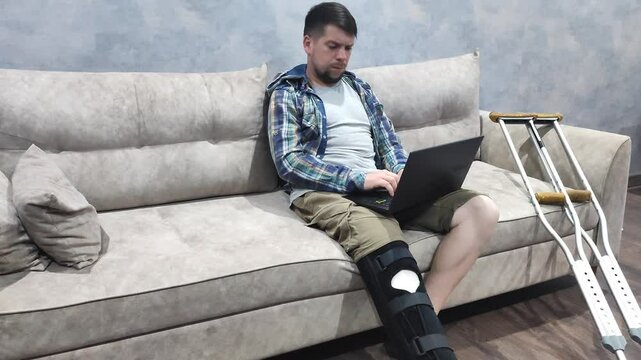 man with orthosis on broken leg sits on sofa and works on laptop. Work from home during illness and recovery, recovery after leg surgery