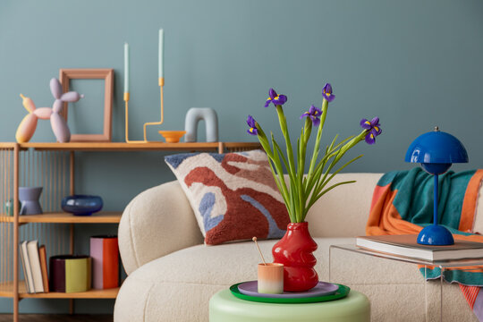 Modern living room with playful decor accents. A cozy sofa adorned with a patterned cushion, vibrant vases, and a blue lamp, complemented by fresh irises in a bold red vase.