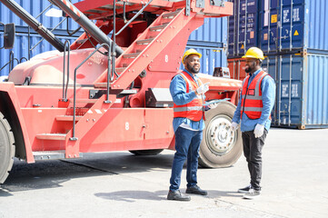 Two African American logistic workers wearing reflective vests and white helmets talk about logistics operations at shipping container yard. Transportation import and export logistic industry concept.