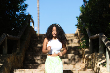 Naklejka premium Woman, young, beautiful, Latin, brunette with curly hair, dressed in sportswear, listening to music with headphones while jogging down a staircase in the park. health and sport concept.