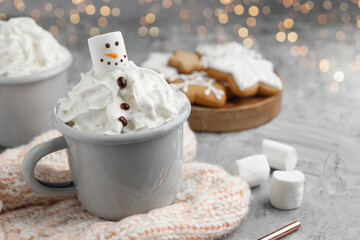 Funny snowman made with marshmallow and whipped cream in cup of hot drink served on grey background against Christmas lights, closeup