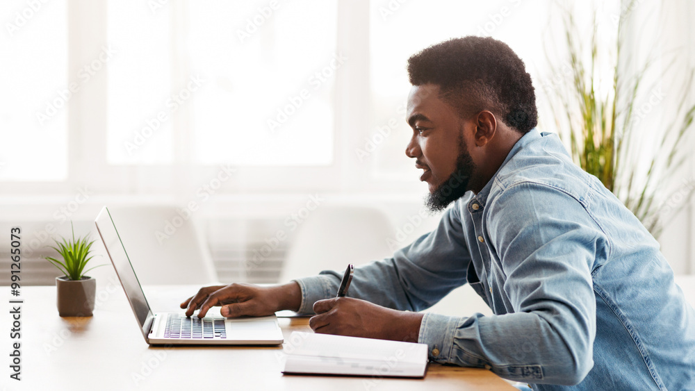 Poster young african american employee using laptop and taking notes while working in modern office, side v