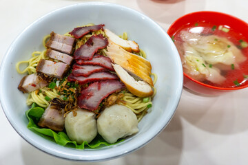 a bowl of noodles served with fish ball, chicken, and pork meat on white bowl and wonton soup on red bowl