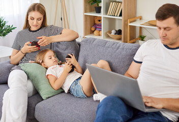 Family at home using laptop and smartphones sitting on sofa, entertaining online obsessed with...