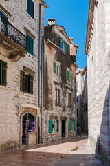 Strolling Kotor's narrow cobblestone streets, visitors admire weathered stone buildings with vibrant green shutters, embracing the local atmosphere under a clear blue sky.