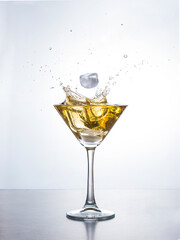 Yellow cocktail in martini glass with ice cube splashing into liquid against white background. Martini cocktail with splash on white background