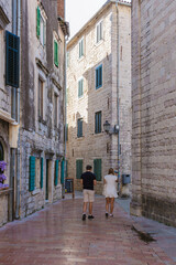 A couple leisurely walks hand in hand through the narrow, sunlit streets of Kotor, surrounded by historic stone buildings and vibrant greenery, enjoying the serene atmosphere.