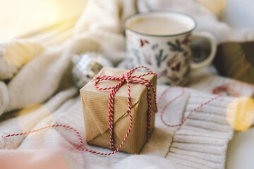 A small gift, carefully wrapped in brown paper and red ribbon, rests on a soft sweater surrounded by soft holiday lights, creating a warm holiday atmosphere.