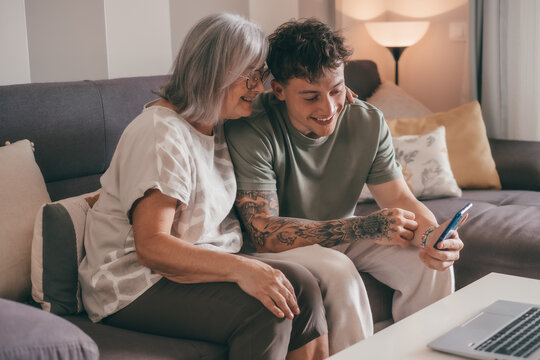 A 70-year-old woman and a young 20-year-old grandson look at the boy's cell phone together while sitting on the couch at home. Friendship and affection in a multigenerational family