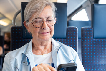 An elderly woman enjoys the train ride sitting with her cell phone in her hand while typing a message, a smiling elderly woman savors the time spent on the journey by reading © luciano