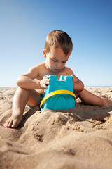 A boy plays in the sand with a blue bucket to build a sand castle.