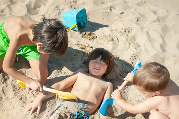 Two children burying their brother in the sand with shovels. © OR MIXphoto