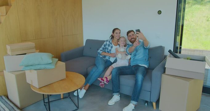 Beautiful family making memorable selfies on smartphone while relaxing, sitting between cardboard boxes after house move.
