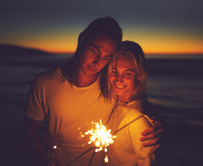 Night, beach and couple with sparklers for holiday, vacation and celebrate new year together. Smile, man and woman with fireworks by sea for romantic date, travel and hug for love outdoor at sunset